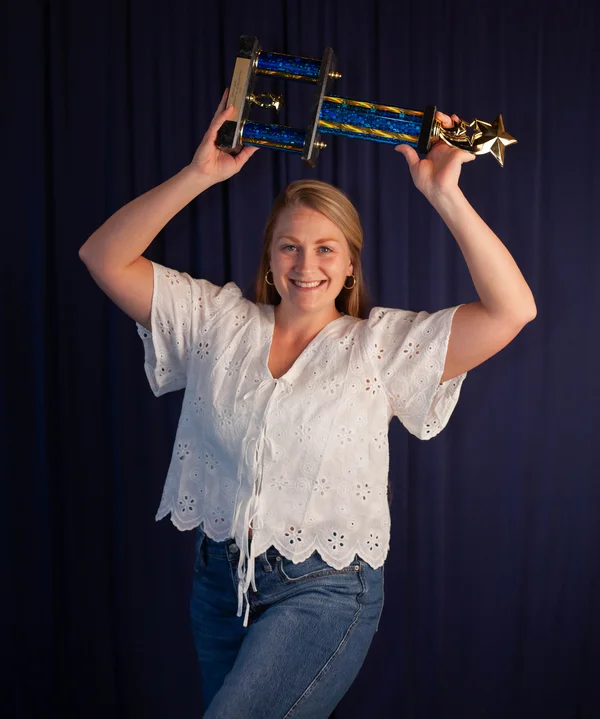 Smiling woman in a white blouse holding a blue and gold trophy above her head against a dark background.