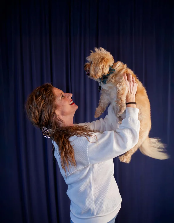 Smiling woman in a white sweater lifting a small fluffy dog wearing a blue bow tie against a dark blue curtain background.
