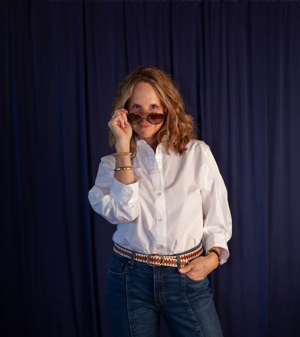 Woman with shoulder-length wavy hair wearing a white button-up shirt and blue jeans, holding sunglasses down to look over them with a smile.