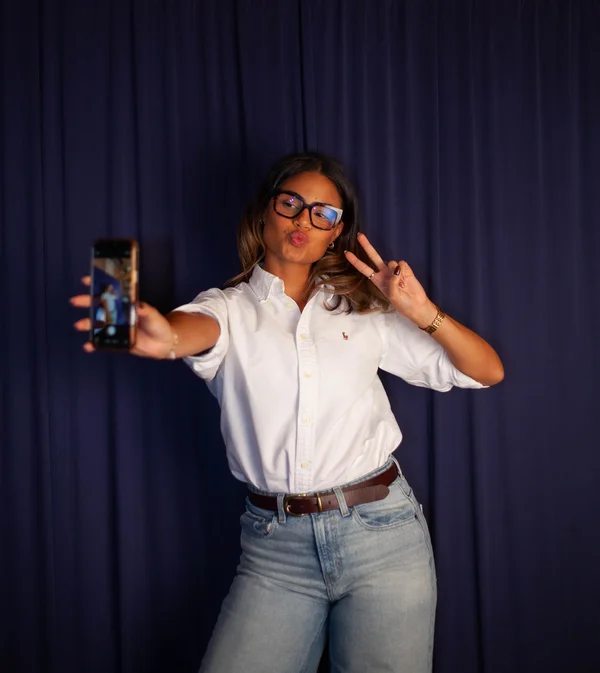 Woman wearing glasses and a white shirt making a peace sign while taking a selfie with a smartphone against a dark curtain backdrop.