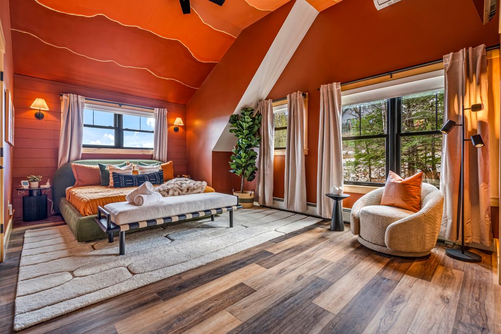 Cozy bedroom with burnt orange walls and ceiling, a low bed with orange bedding, a bench with towels, beige rug, a large potted plant, and a curved armchair beside windows with beige curtains.