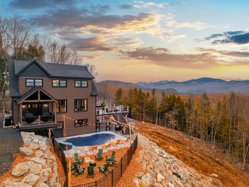 Modern brown house on a rocky hillside overlooking a forest with autumn foliage and distant mountains at sunset.
