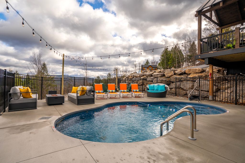 Outdoor swimming pool with orange lounge chairs, cushioned seating, string lights, and a rocky hillside under a cloudy sky.