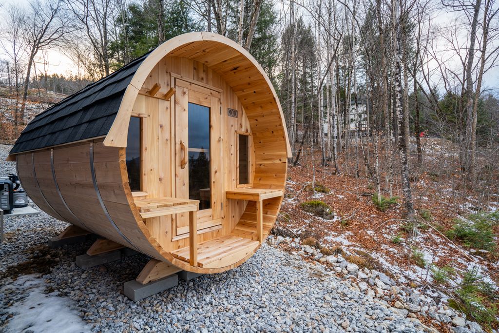 Wooden barrel-shaped sauna cabin with a small porch set on concrete blocks in a forested area with some snow on the ground.