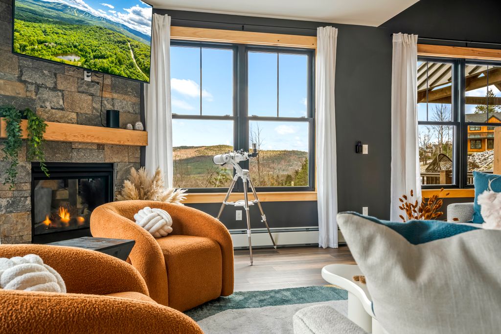 Cozy living room with orange chairs, a fireplace, a mounted TV, large windows showing a scenic mountain view, and a telescope on a tripod.