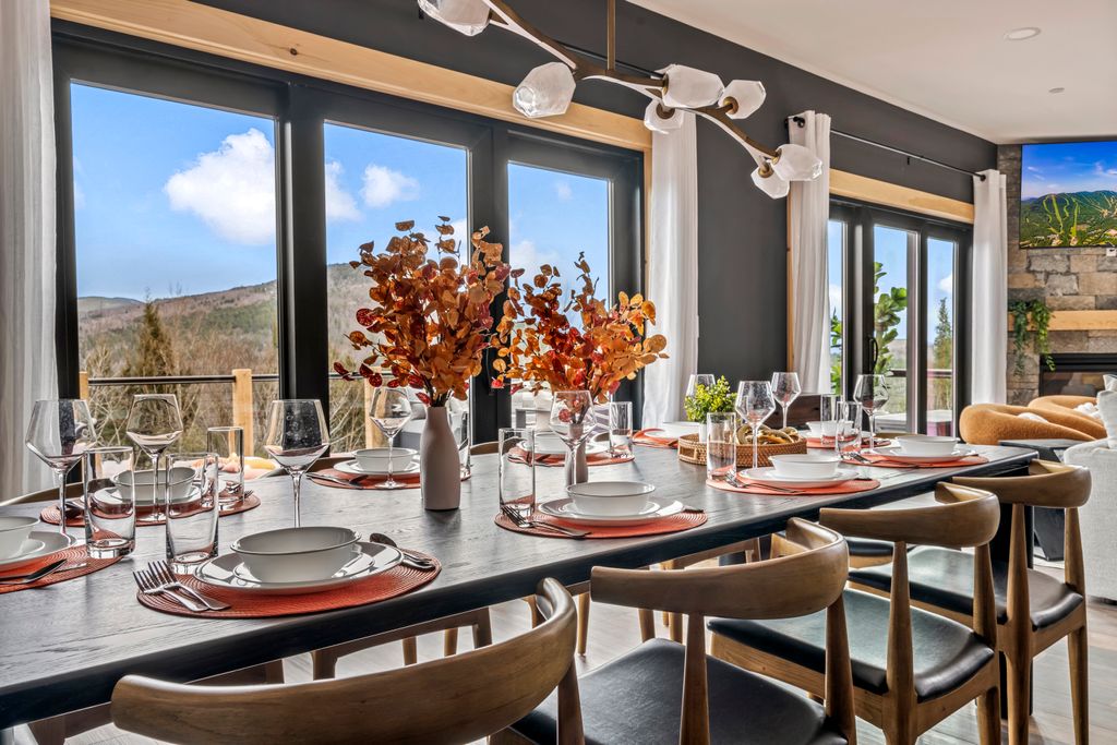 Modern dining table set with white plates, wine glasses, and autumnal floral centerpieces by large windows showing a mountain view.