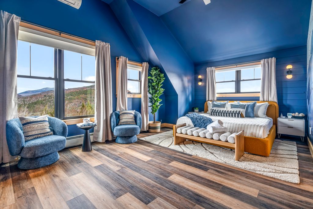 Bright bedroom with blue walls and ceiling, a large window showing a mountain view, two blue upholstered chairs, a wooden bed with white bedding and multiple pillows, and a wooden bench at the foot of the bed holding rolled towels.