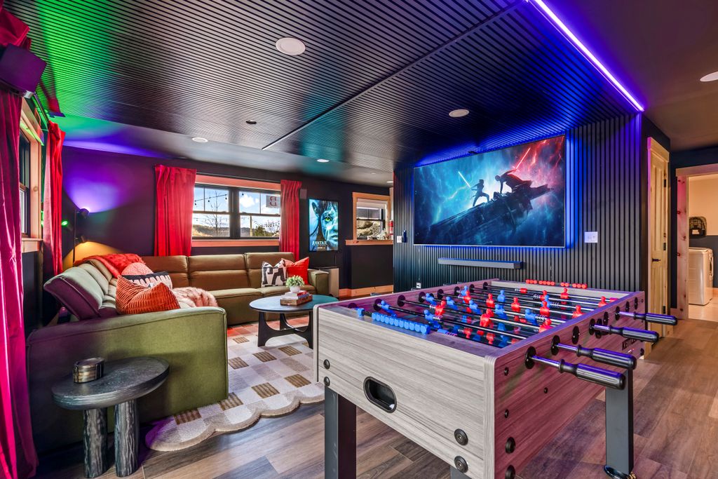 Modern game room with foosball table, large sectional sofa with red pillows, and a wall-mounted TV showing a Star Wars duel scene, illuminated by colorful LED lights.