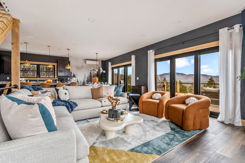 Modern living room with light gray sectional sofa, two orange armchairs, abstract area rug, and large glass doors revealing mountain views.