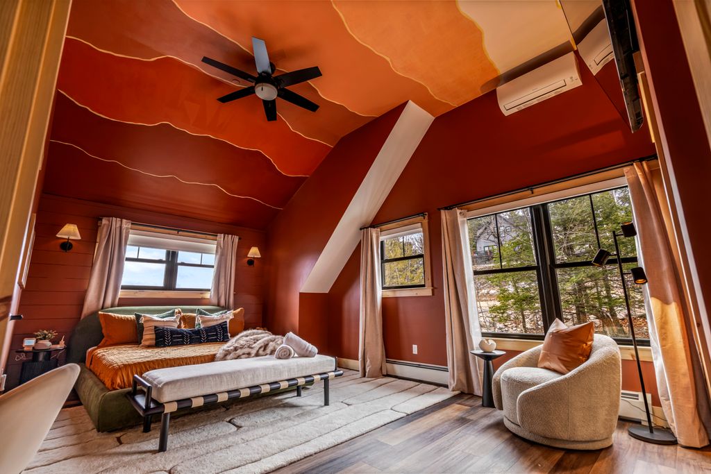 Cozy bedroom with warm-toned walls and ceiling, a bed with orange bedding and multiple pillows, a bench at the foot of the bed, and a white armchair near large windows overlooking trees.