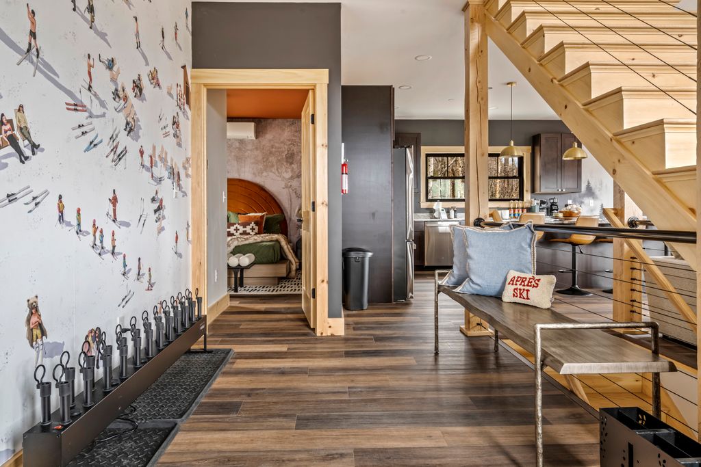 Modern ski lodge interior with wooden flooring, a bench with pillows including one labeled 'Après Ski,' open kitchen, visible bedroom, and ski boot warmers along a wall with ski-themed mural.