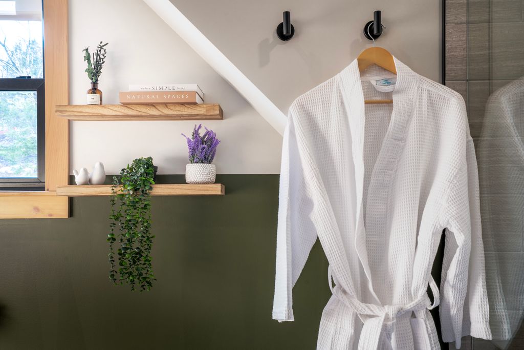 White waffle-textured bathrobe hanging on a wooden hanger on a wall with two black hooks next to wooden shelves holding plants and books.