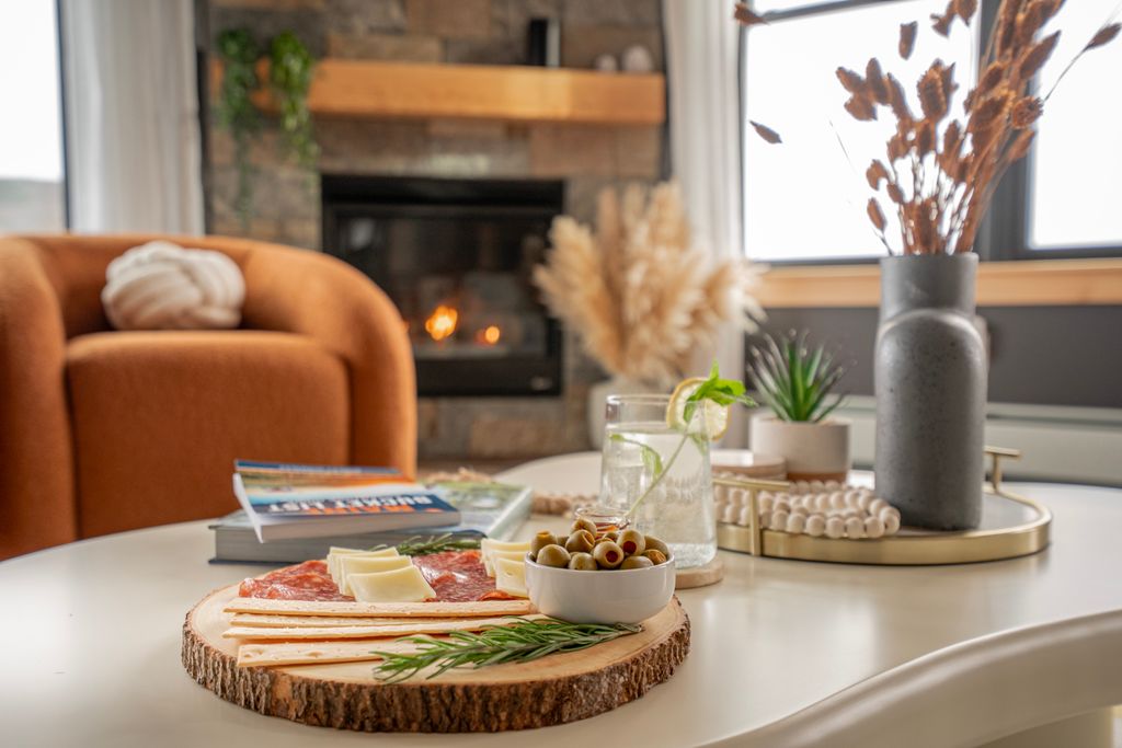 Wooden platter with cheese, salami, breadsticks, and a bowl of olives on a white table in a cozy living room with fireplace and orange armchair.