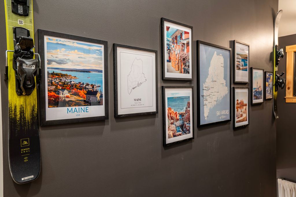 Wall displaying framed pictures and maps of Maine along with a pair of yellow and black skis mounted vertically.