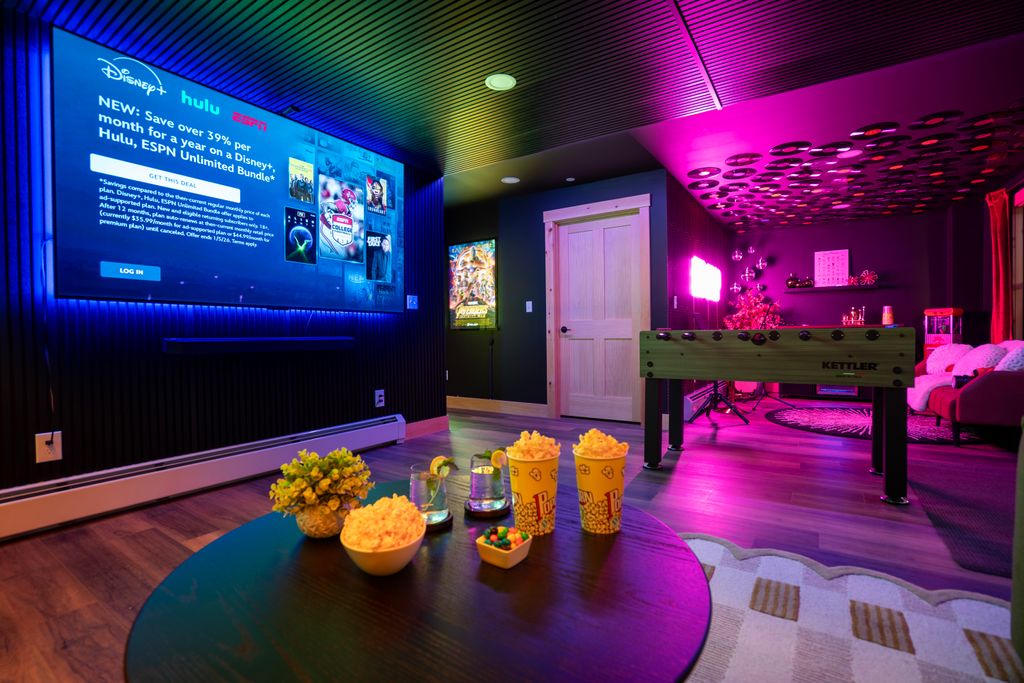 Cozy entertainment room with a large TV screen displaying streaming service offers, a foosball table, colorful ambient lighting, and snacks on a round table.