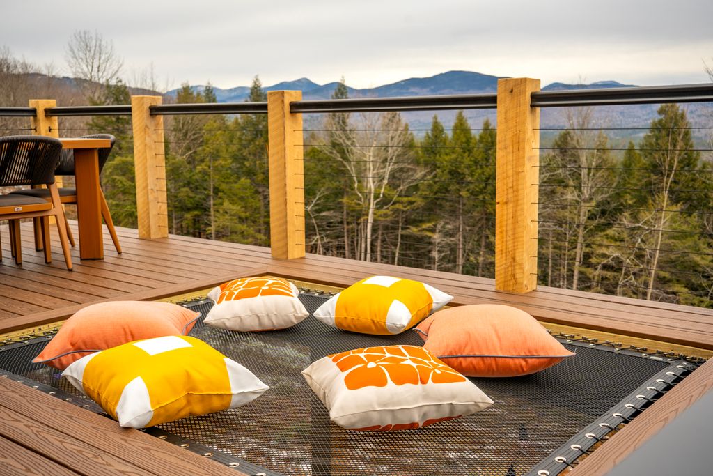 Outdoor wooden deck with a built-in trampoline holding five colorful cushions, overlooking trees and distant mountains.