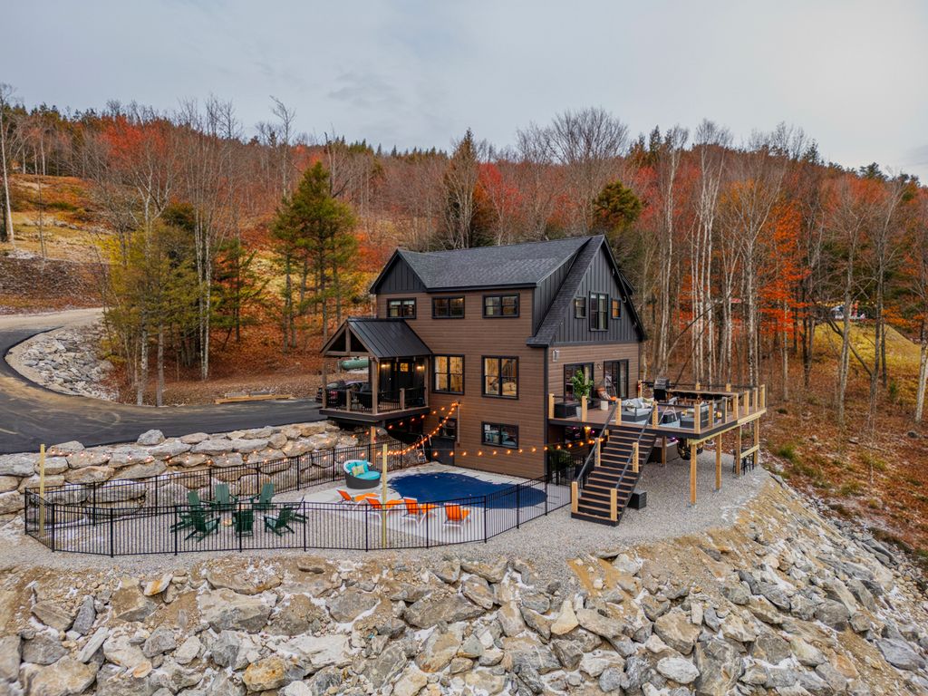 Modern two-story house with a wraparound deck and fenced outdoor pool area surrounded by rocky terrain and autumn trees.