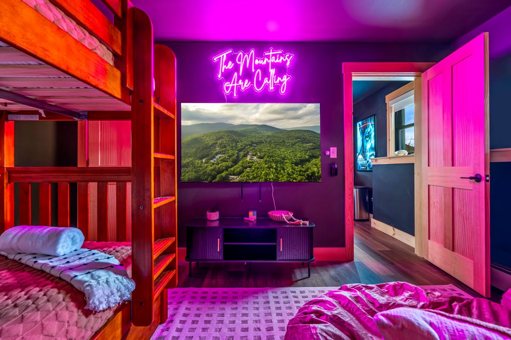 Room with a wooden bunk bed, a wall-mounted TV showing a forested mountain landscape, a neon sign that reads 'The Mountains Are Calling,' and an open wooden door leading to another room.