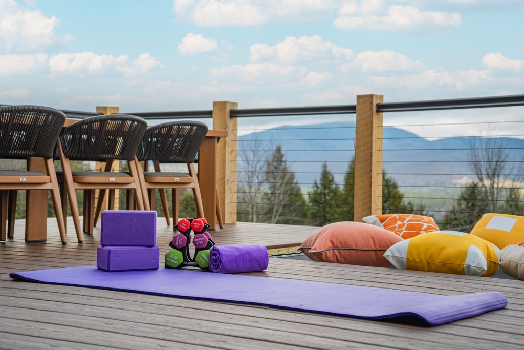 Purple yoga mat with blocks, towel, and dumbbells on a wooden deck overlooking mountains, with colorful cushions and chairs nearby.
