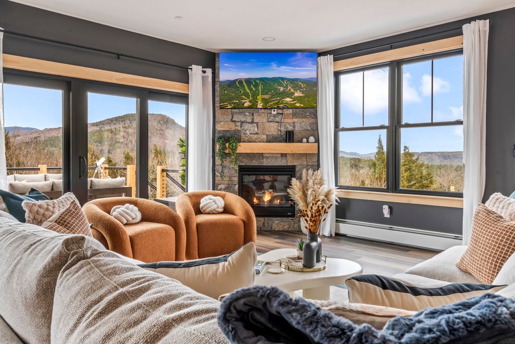 Cozy living room with beige sectional sofa, two brown armchairs, stone fireplace, mounted TV, and large windows showing a mountain view.
