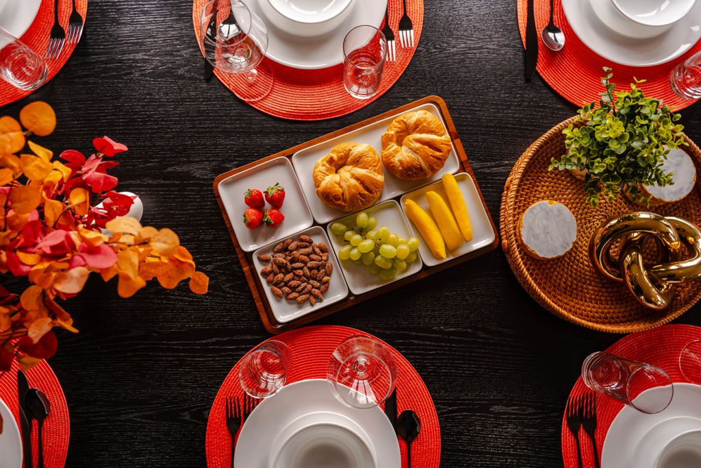 Dining table set with white dishes on red placemats and a wooden tray with croissants, strawberries, grapes, sliced mango, and almonds in small white plates.