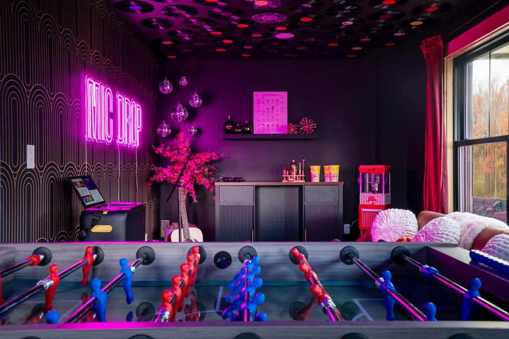 Recreational room with foosball table in foreground, neon MIC DROP sign, pink flowers, guitar, popcorn machine, and large window with red curtains.