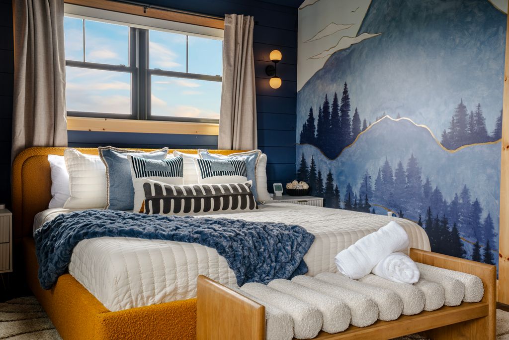 Cozy bedroom with mustard yellow bedframe, white and patterned pillows, blue throw blanket, and mountain mural painted on the wall.