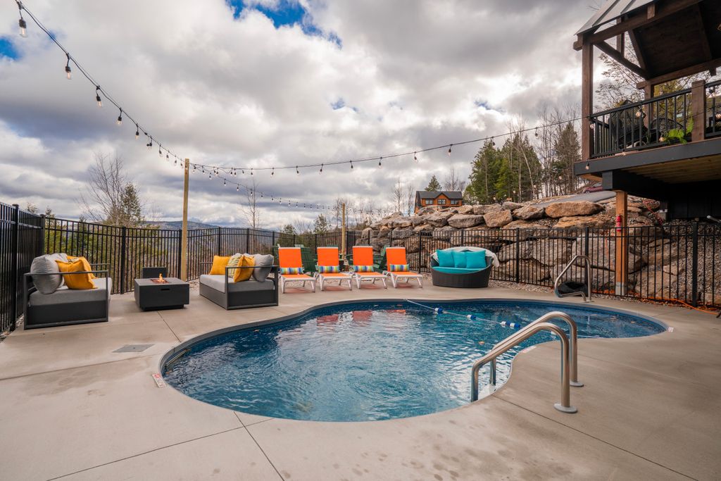 Outdoor kidney-shaped pool with lounge chairs featuring orange cushions, gray seating with yellow pillows, a round teal cushioned chair, and string lights overhead against a cloudy sky.