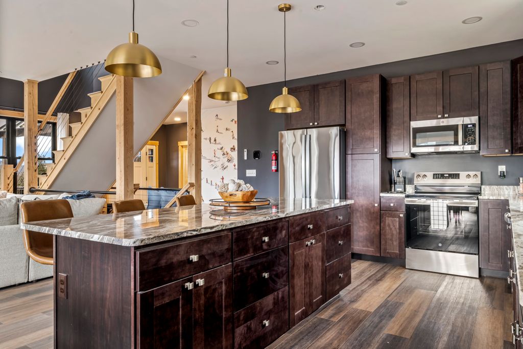 Modern kitchen with dark wood cabinetry, stainless steel appliances, a large marble island with brown leather chairs, and three gold pendant lights.
