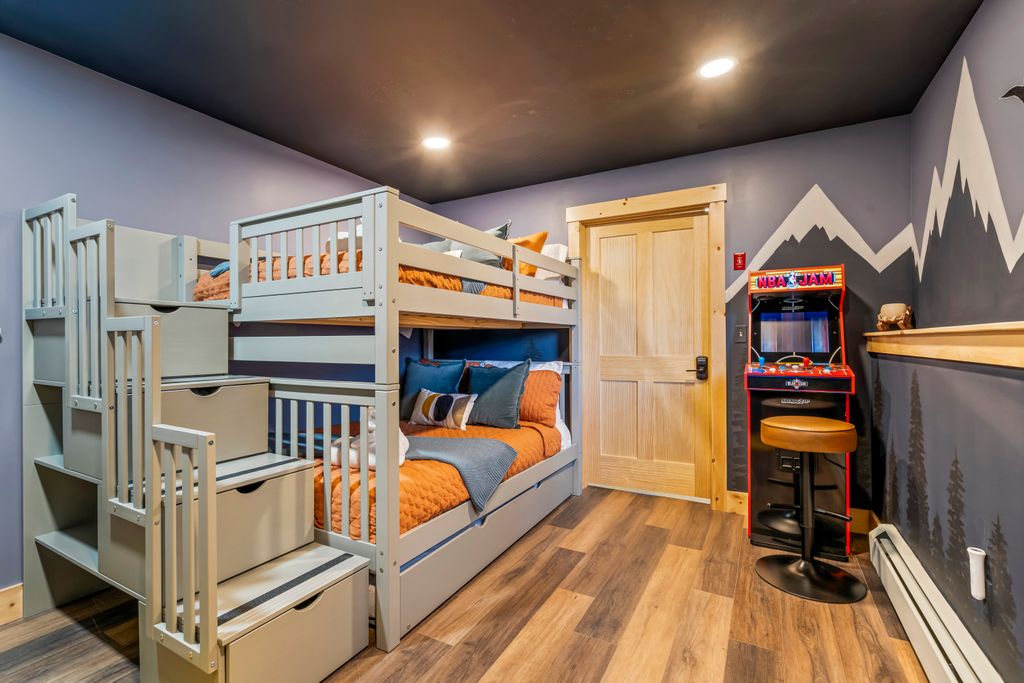 Modern bedroom with gray bunk bed featuring orange bedding, wooden floor, mountain mural on gray walls, and a vintage NBA Jam arcade machine with stool.