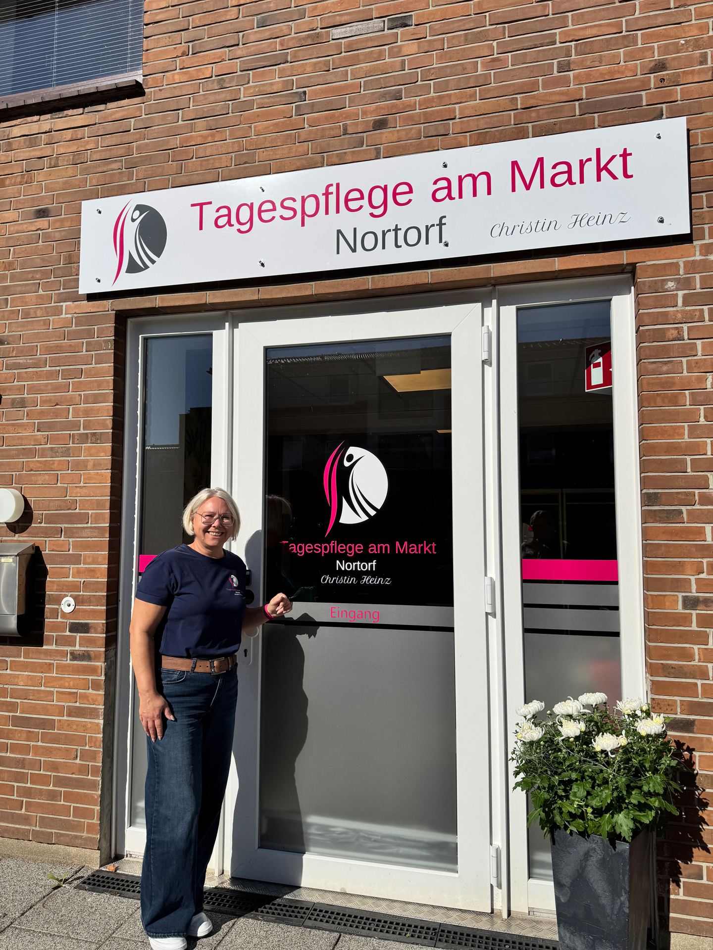 Smiling woman with glasses standing at the entrance door of Tagespflege am Markt Nortorf building with white flowers in a black planter nearby.