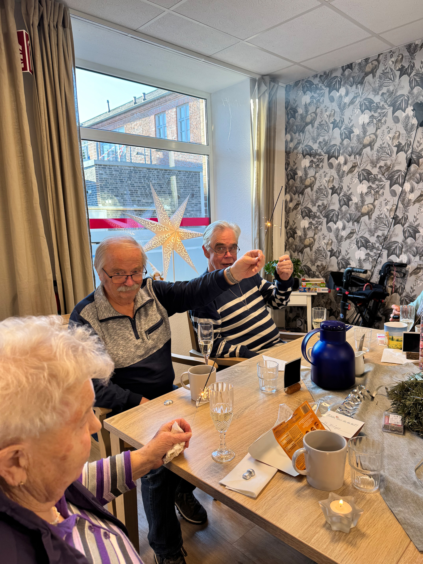 Three elderly people sitting around a table inside a room holding lit sparklers, with drinks and decorations on the table.