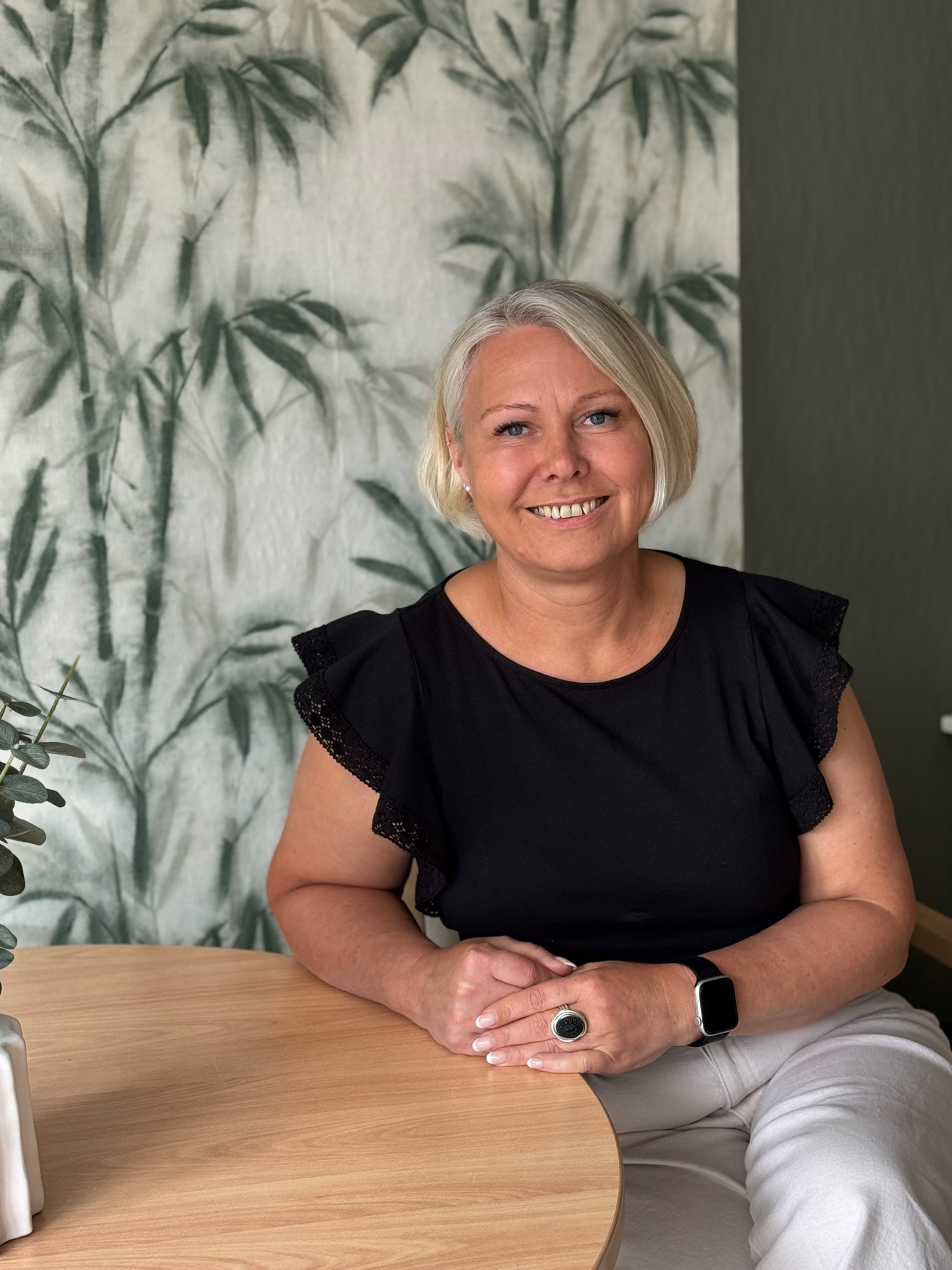 Smiling woman with short blonde hair wearing a black top and white pants sitting at a wooden table with a plant and bamboo-patterned background.