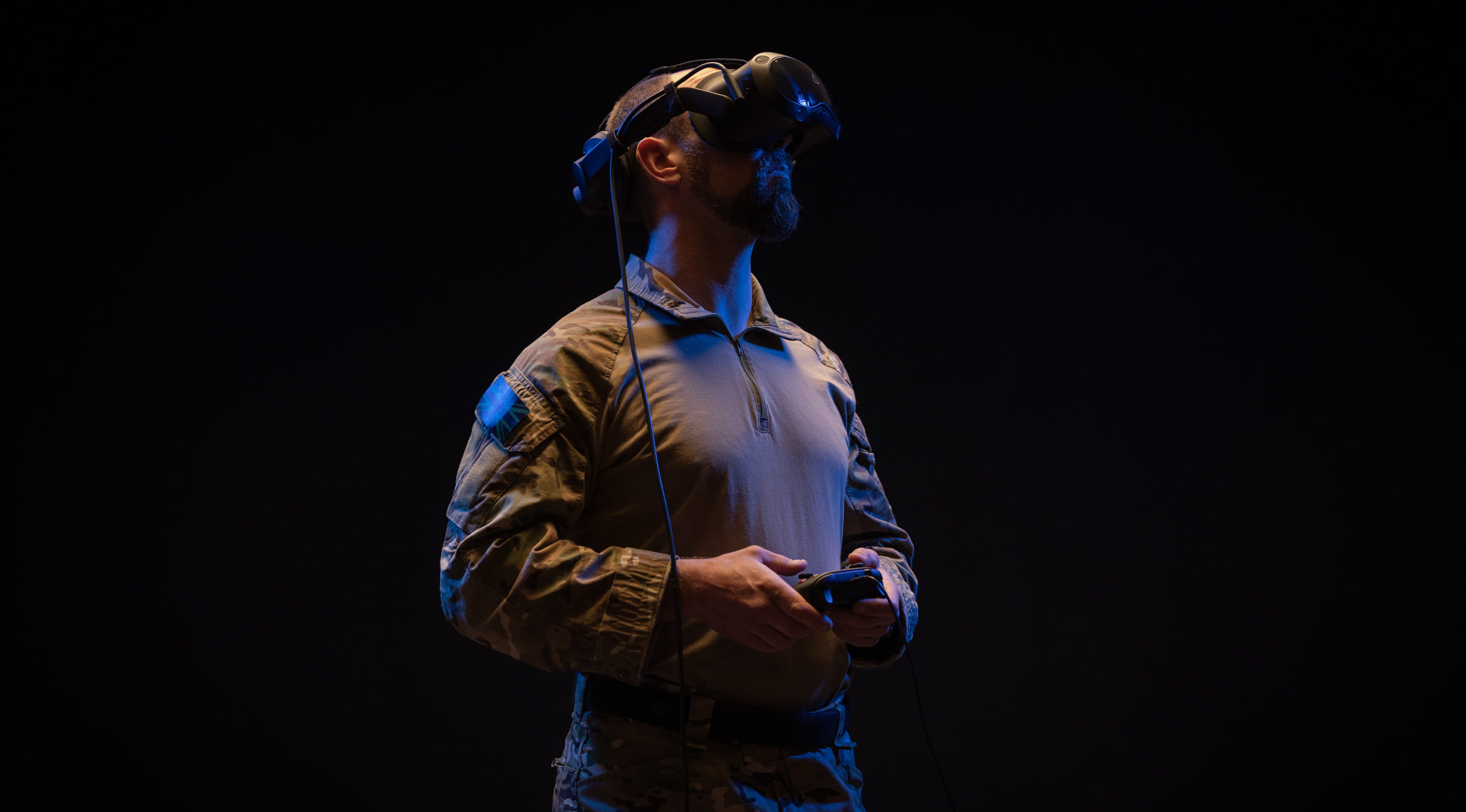 Man in military camouflage uniform using a virtual reality headset and holding a controller against a dark background.