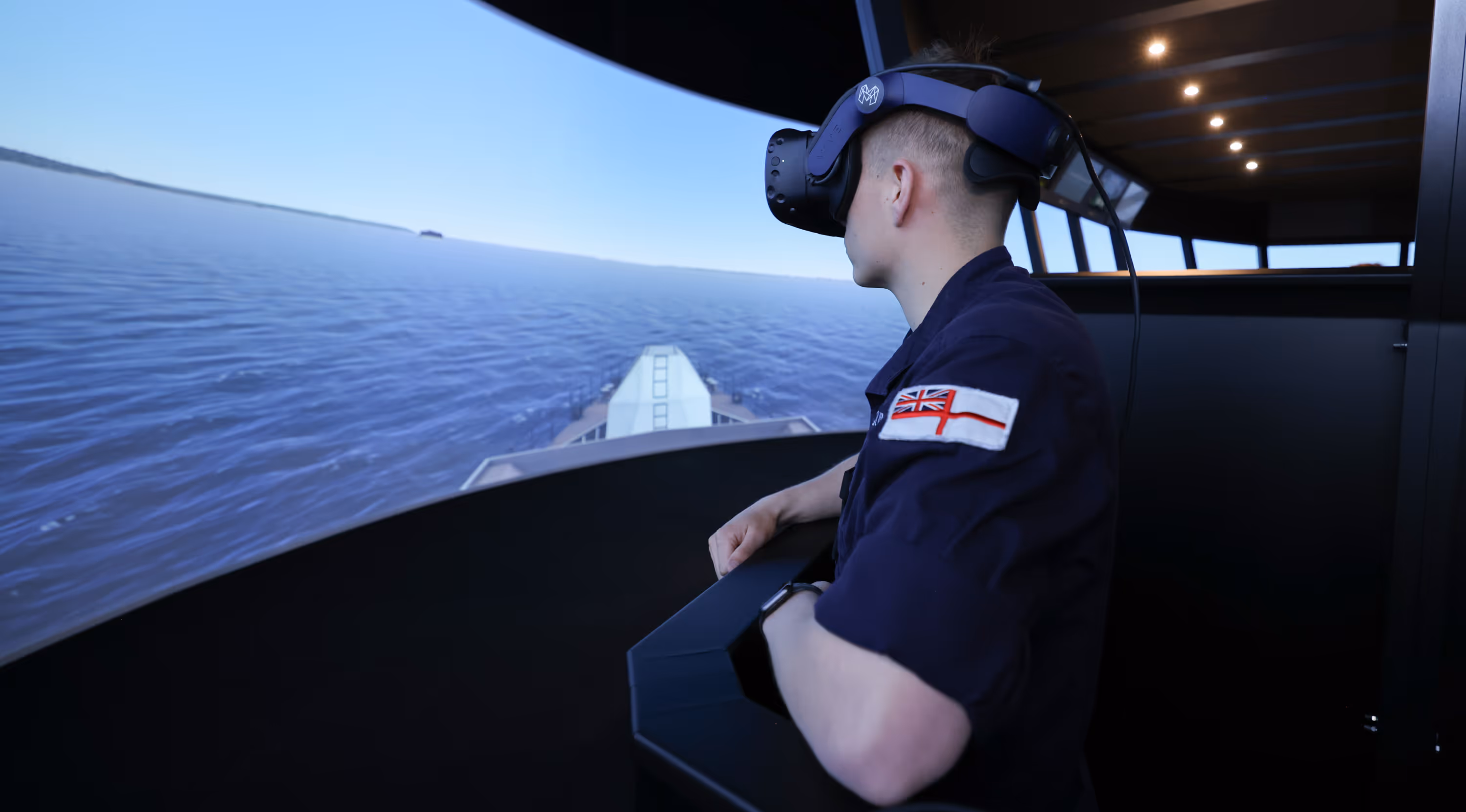 Person wearing virtual reality headset inside a simulator, overlooking a digital ocean and ship's bow view.