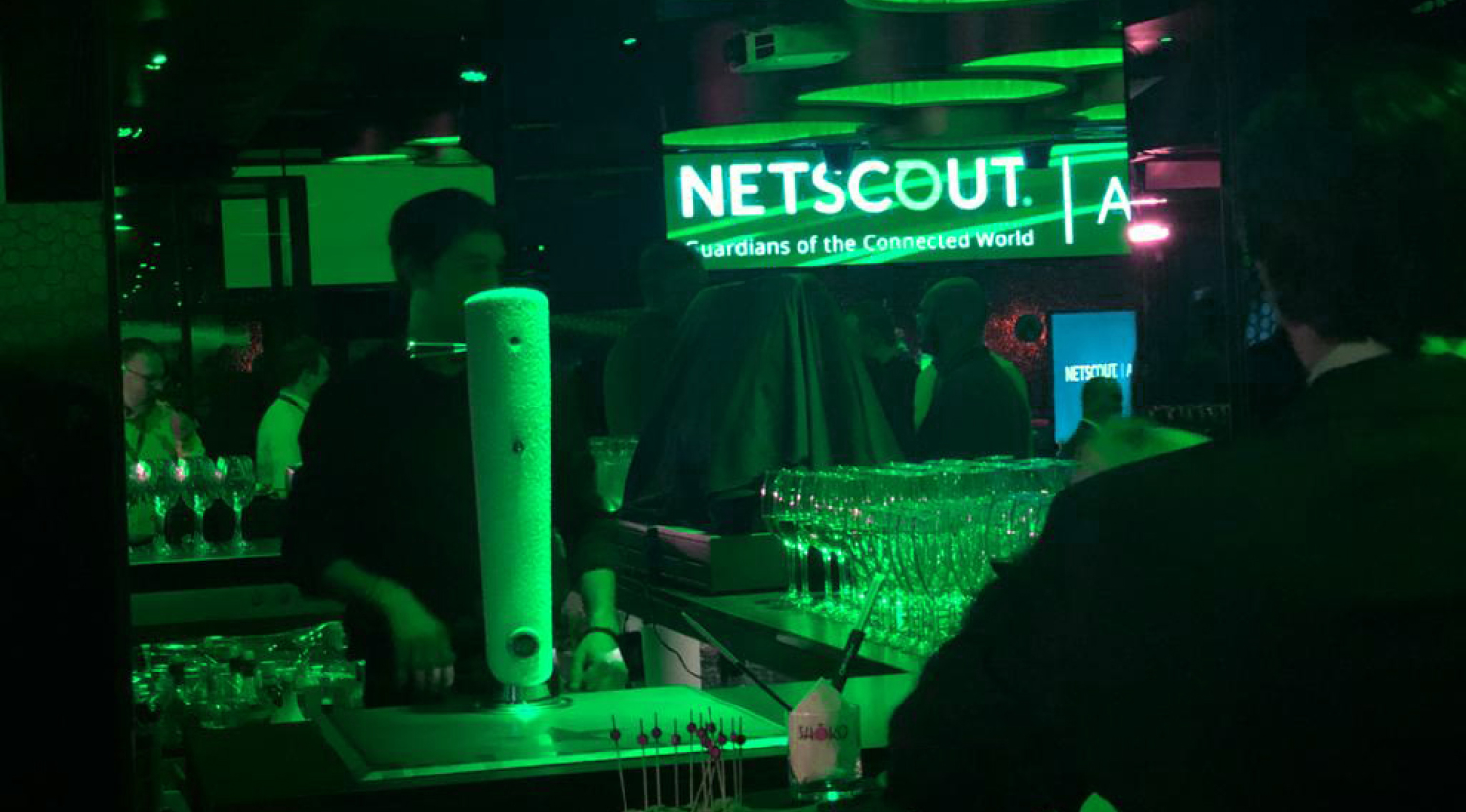 Dim green-lit bar scene with people, glassware, and a illuminated NETSCOUT sign reading 'Guardians of the Connected World'.