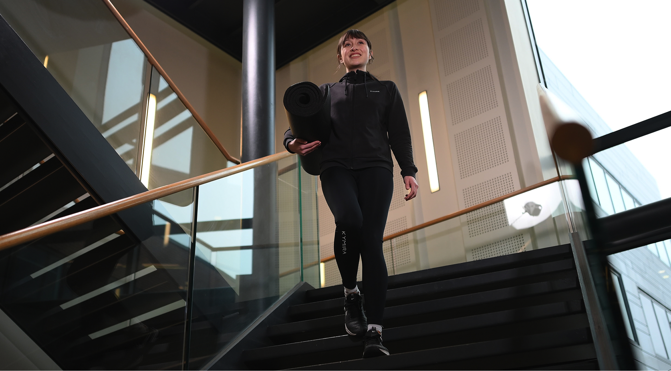 An athlete walking down the stairs in KYMIRA recovery wear, with a yoga mat under her arm