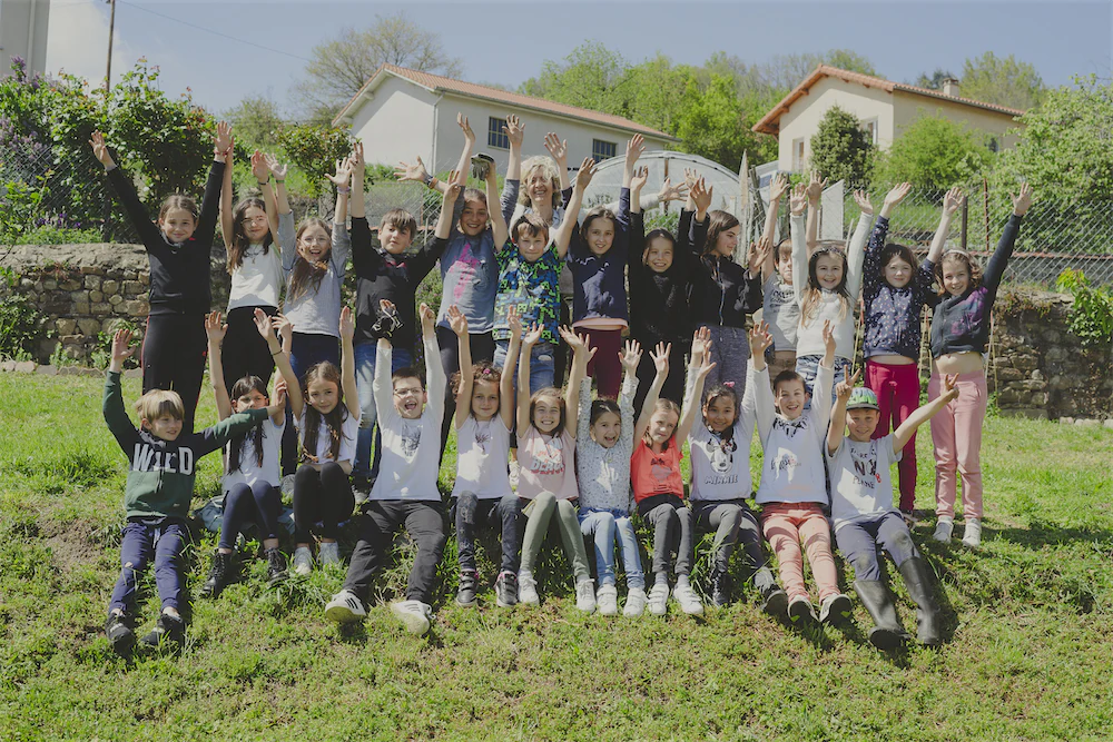 Groupe d'enfants assis et debout sur l'herbe avec les bras levés, avec des maisons et un ciel bleu en arrière-plan.