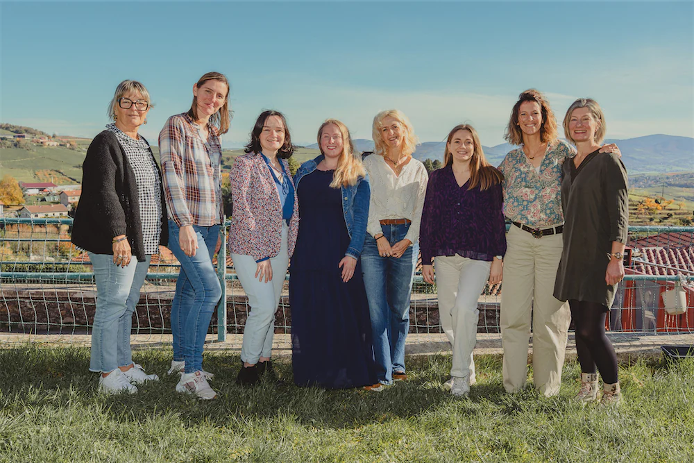 Groupe de huit femmes souriantes travaillant dans l'école de Saint-Joseph debout sur l'herbe avec un paysage de collines et maisons en arrière-plan sous un ciel bleu clair.