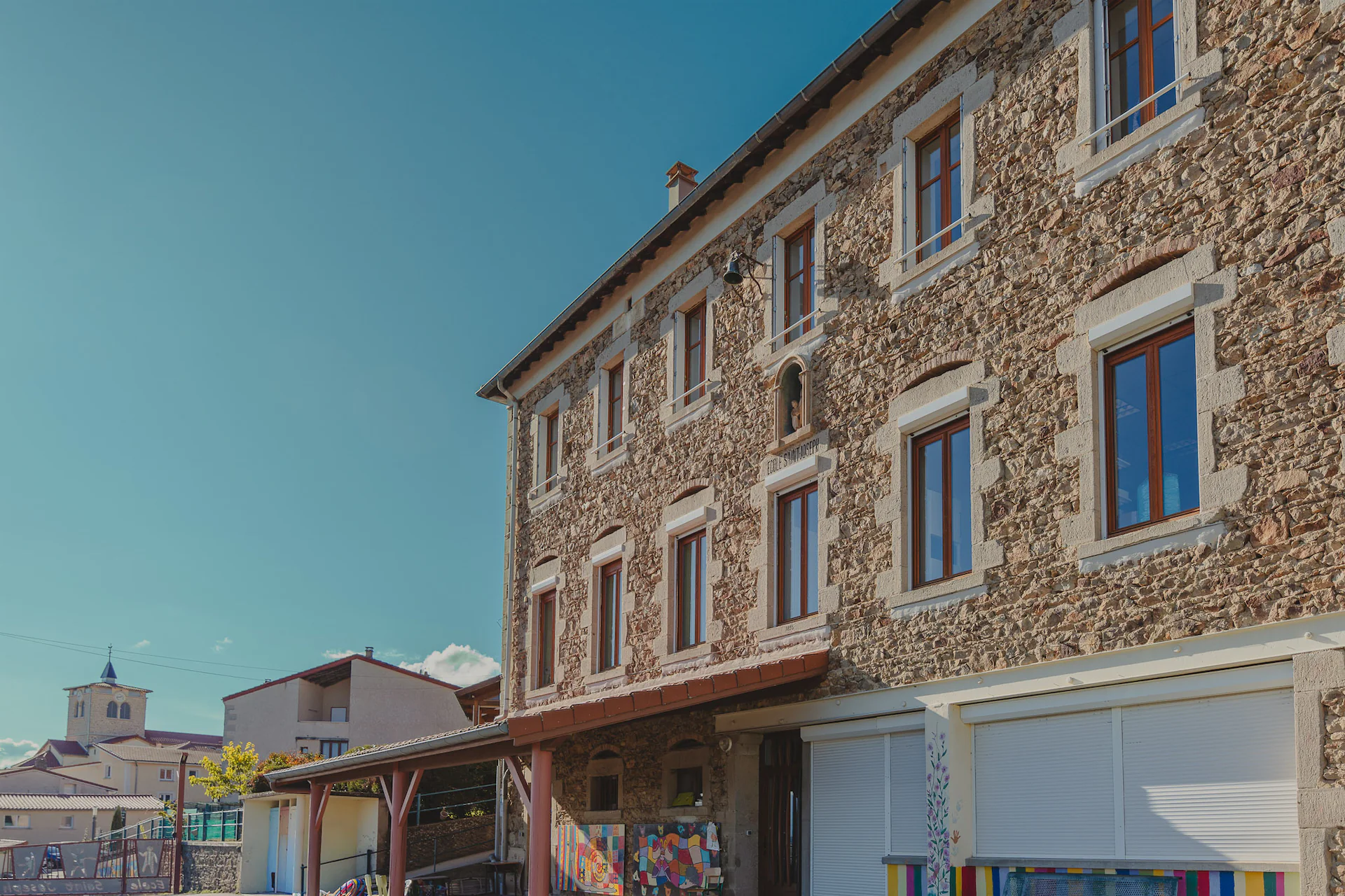 Façade en pierre de l'école Saint-Joseph avec plusieurs fenêtres, au ciel bleu, et une église en arrière-plan.