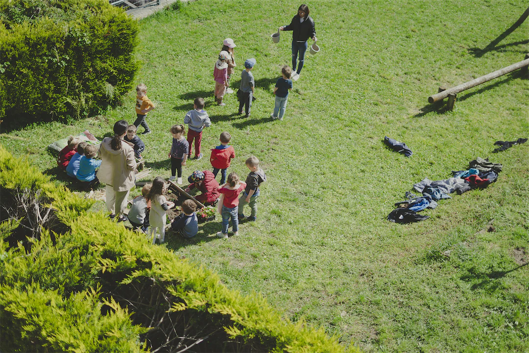 Un groupe d'enfants assis et debout sur une pelouse verte, entourés d'arbustes, avec deux adultes supervisant une activité en plein air.