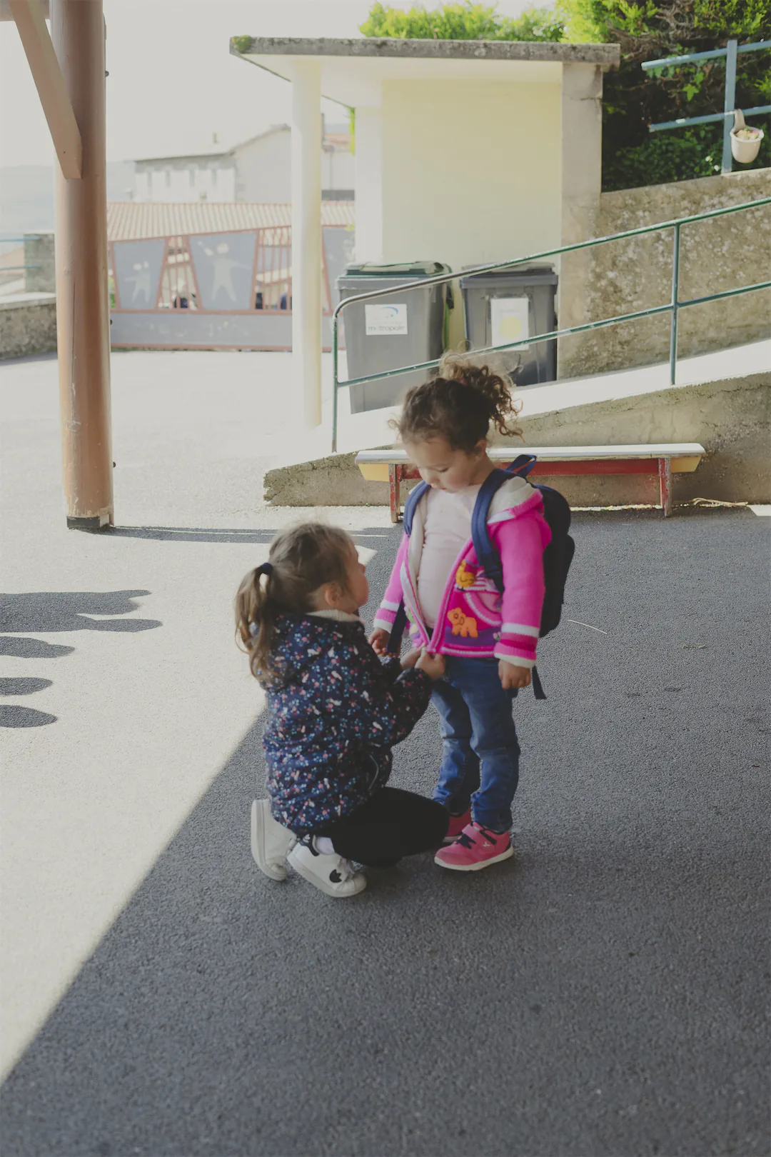 Deux jeunes filles se tiennent dehors, l'une est accroupie en train d'aider l'autre avec sa veste rose et son sac à dos bleu.