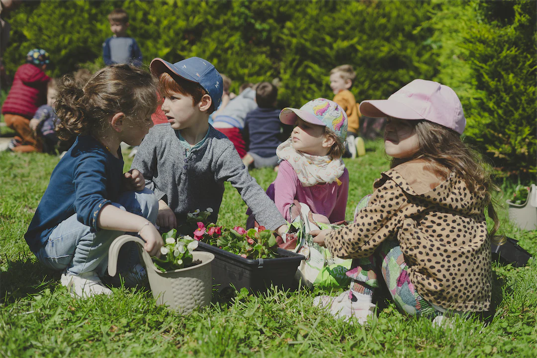 Quatre enfants assis sur l'herbe jardinent ensemble avec des fleurs et des pots dans un espace vert extérieur.