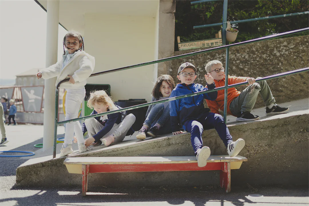 Cinq enfants heureux jouent sur une rampe extérieure en béton, un garçon en veste bleue fait signe de la main.