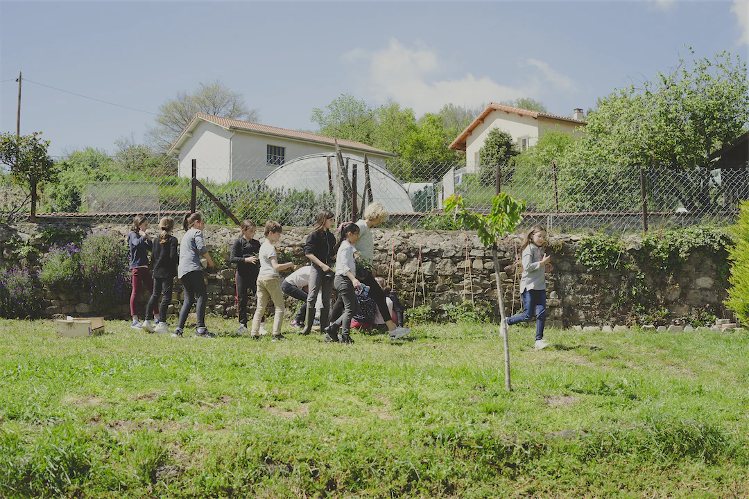 Un groupe d'enfants et un adulte dans un jardin avec herbe, entouré de murs en pierre et de maisons en arrière-plan sous un ciel bleu.