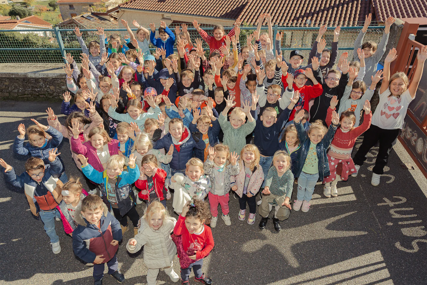 Toute l'équipe et enfants de l'École Saint-Joseph souriants levant les mains en l'air dans une cour extérieure ensoleillée.