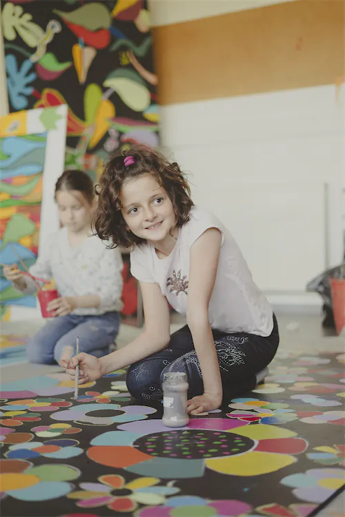 Deux filles assises par terre en train de peindre une grande toile colorée avec des formes fleuries.