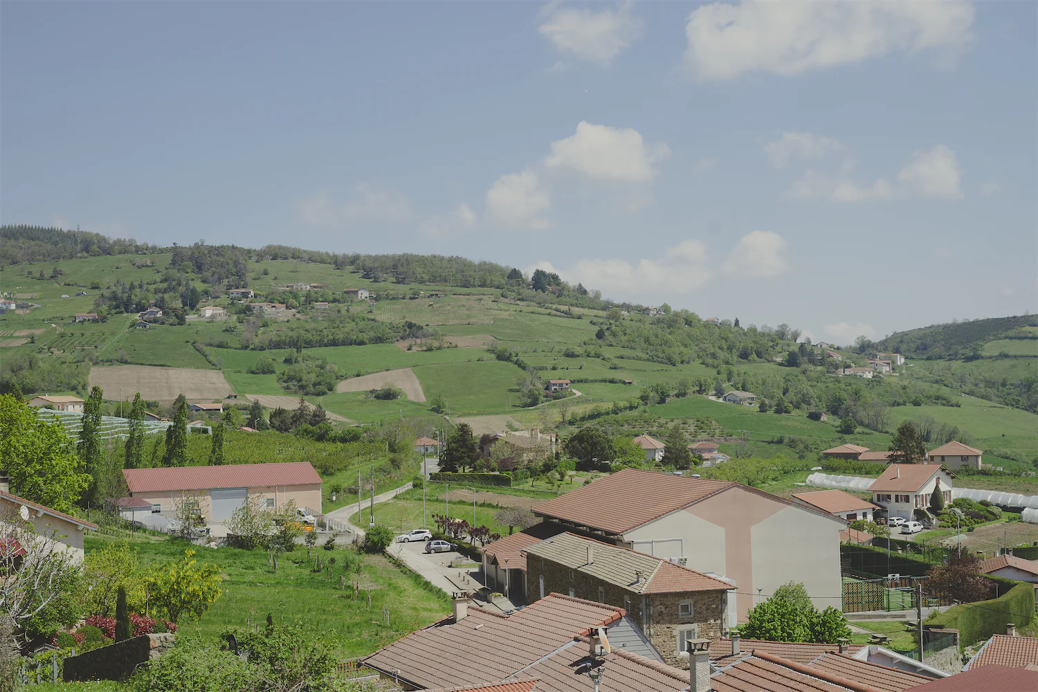 Paysage rural sur la commune de Saint-Romain-en-Jarez avec maisons aux toits rouges, champs verts et collines sous un ciel bleu clair avec quelques nuages.