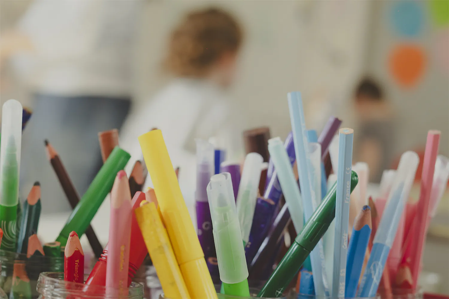 Une collection de crayons de couleur et marqueurs multicolores dans des pots en verre, avec un fond flou d'enfants en classe.