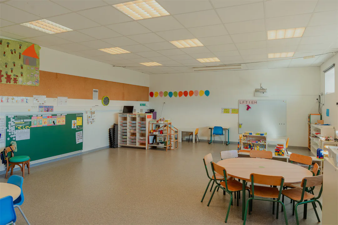 Salle de classe pour jeunes enfants avec tables rondes, chaises colorées, tableau blanc, et espace de rangement équipé de jouets et matériel pédagogique.